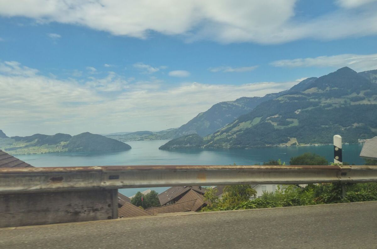 Een panoramisch uitzicht op een groot meer omringd door groene bergen en heuvels onder een deels bewolkte blauwe lucht. Op de voorgrond is een vangrail van een weg zichtbaar, met daaronder de daken van enkele huizen en groen struikgewas. Het meer heeft een helderblauwe kleur en in de verte zijn kleine eilandjes of landtongen te zien.