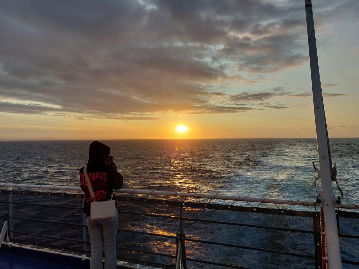 Een persoon, gekleed in een hoodie en jeans, staat met de rug naar de camera gericht op het dek van een schip. De persoon kijkt uit over de open zee waar de zon laag aan de horizon staat. De zonsondergang kleurt de lucht en het water met oranje en gele tinten, met reflecties op de golven. De lucht erboven is deels bewolkt. Aan de rechterkant is een deel van een witte scheepsmast zichtbaar en op de voorgrond de donkere reling van het schip.