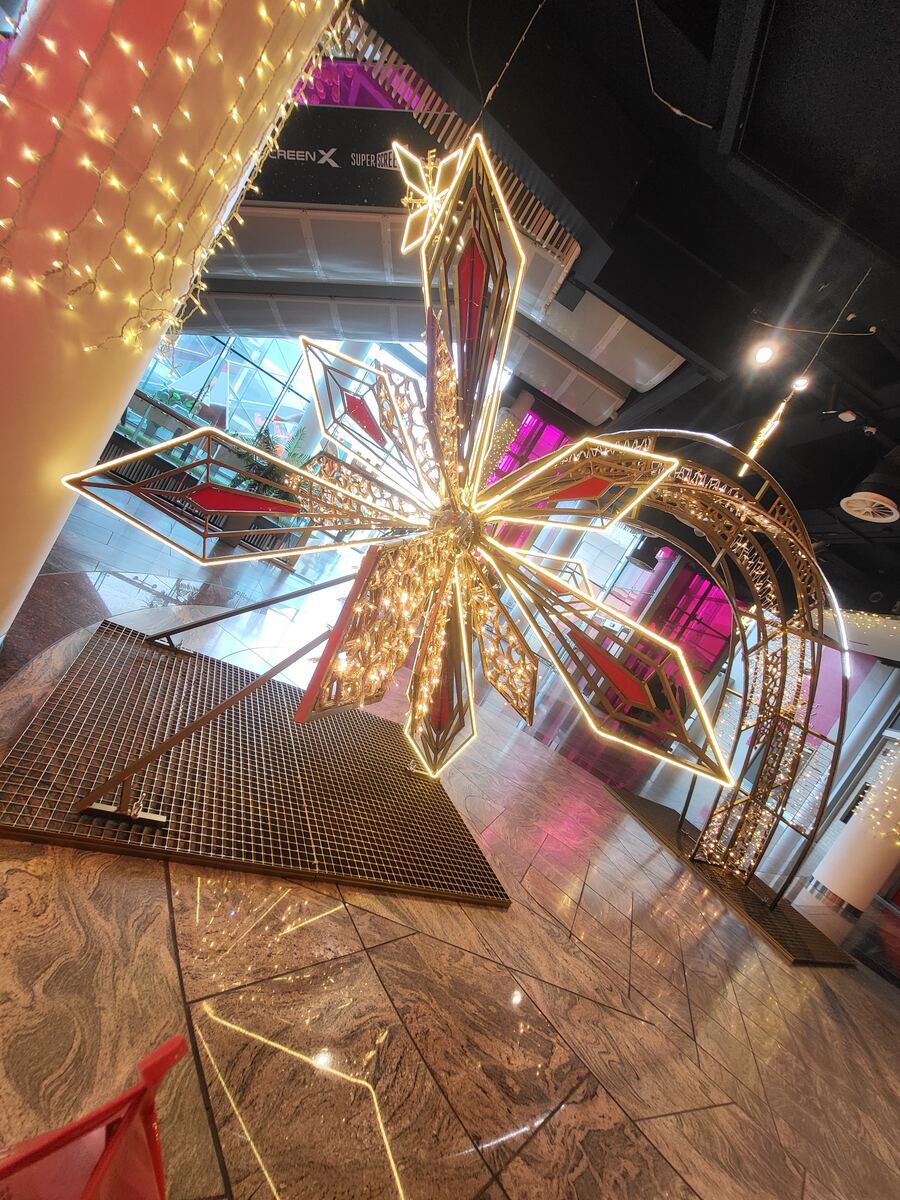Een grote, stervormige decoratie met gouden en rode accenten en veel kleine lampjes hangt in een modern gebouw. De vloer bestaat uit glanzende marmeren tegels. Op de achtergrond zijn glazen wanden en een deel van een reclamebord met 'SCREEN X SUPER REEL' te zien. Er hangen ook andere lichtslingers en decoraties.