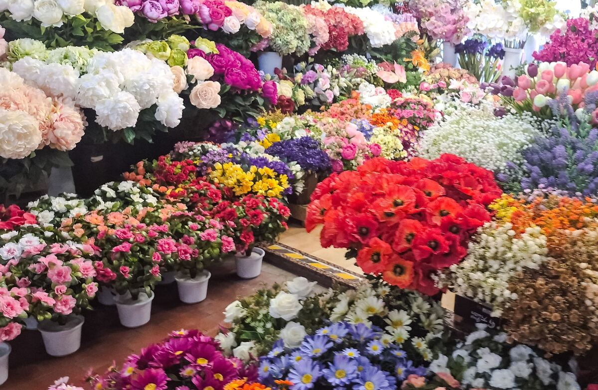 De foto toont een grote, kleurrijke verzameling kunstbloemen. Er zijn bossen helderrode klaprozen, witte hortensia's, roze rozen, gele bloemen, paarse bloemen, wit gipskruid en diverse andere bloemsoorten in verschillende kleuren. De bloemen staan in potten en vazen opgesteld, waardoor een dicht gevuld en levendig beeld ontstaat van een bloemendisplay in een winkel.