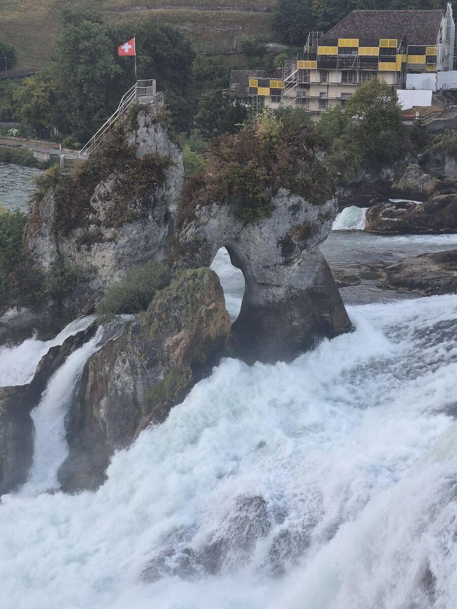 Een verticale foto van de Rijnwaterval met een Zwitserse vlag op een rots in het midden van de waterval, omringd door kolkend water. Op de achtergrond is een gebouw met steigers te zien.