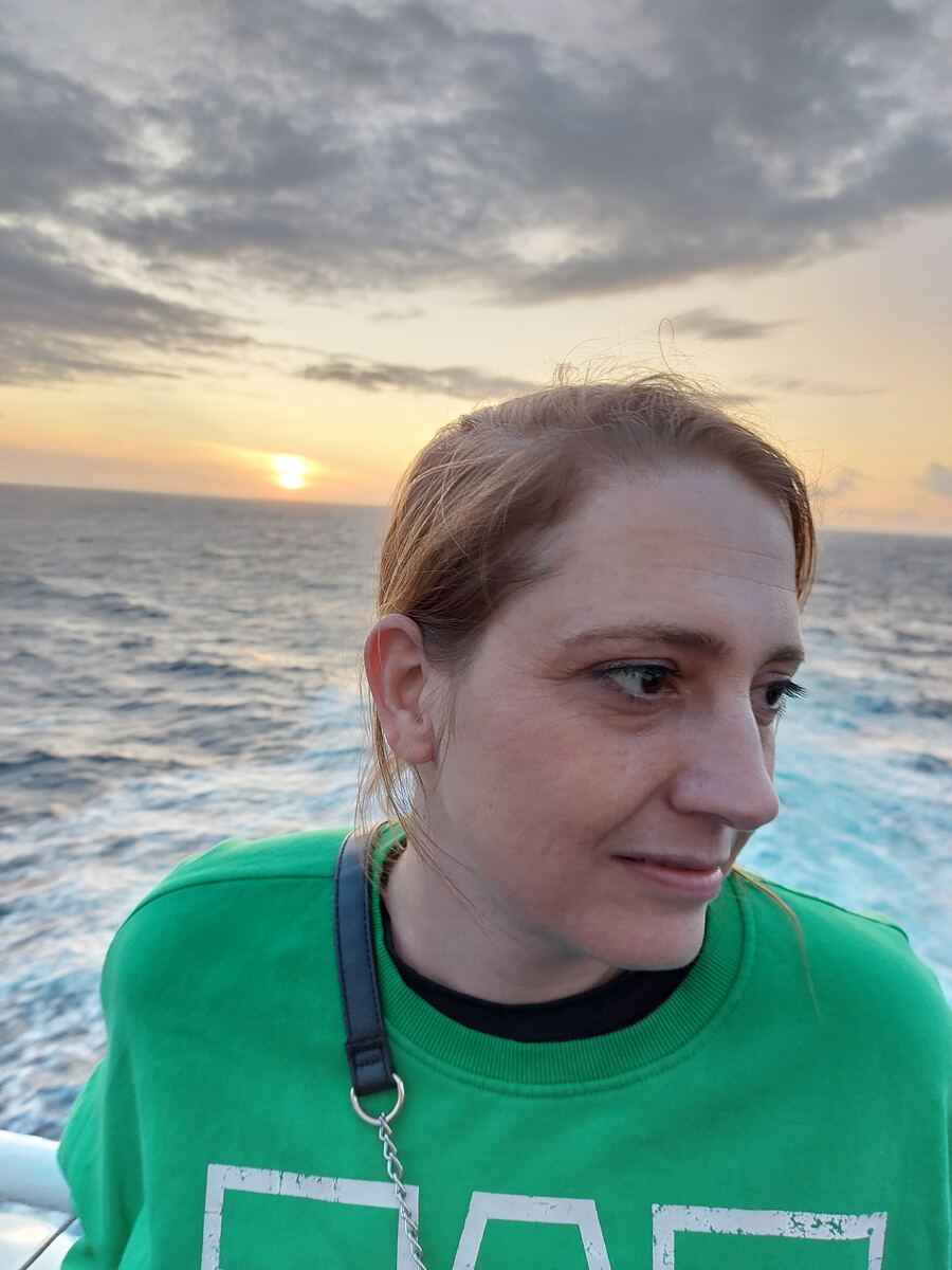Een vrouw in een groene trui kijkt naar rechts, met op de achtergrond de open zee en een ondergaande zon aan de horizon. De lucht is deels bewolkt en de zon reflecteert op het wateroppervlak. Links is een witte reling zichtbaar, wat duidt op een vaartuig.