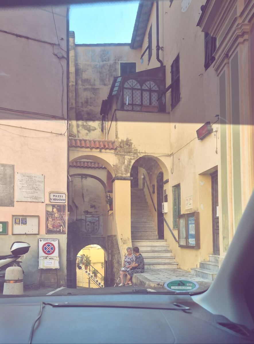 De foto toont een smalle straat in een oudere stad, gezien vanuit een auto. Aan de linkerkant is een lichtroze gebouw met verschillende borden en plaquettes aan de muur. Een van de borden vermeldt 'PIAZZA S SEBASTIANO' en een ander 'MUSEO CIVICO VILLA LUCA'. Er is ook een verkeersbord met een rood kruis op een blauwe achtergrond, wat duidt op een parkeerverbod. Aan de rechterkant staat een geel gebouw met een trap die omhoog leidt. Twee oudere vrouwen zitten op de onderste trede van deze trap. A