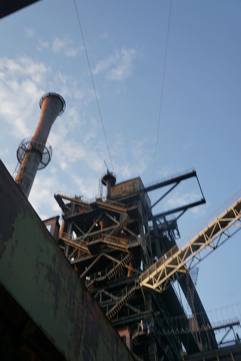 Een foto genomen vanuit een laag standpunt, gericht op een deel van een grote industriële installatie met een schoorsteen en complexe metalen constructies, waaronder trappen en transportbanden. De structuren zijn roestig en tonen tekenen van slijtage. De lucht is blauw met enkele witte wolken.