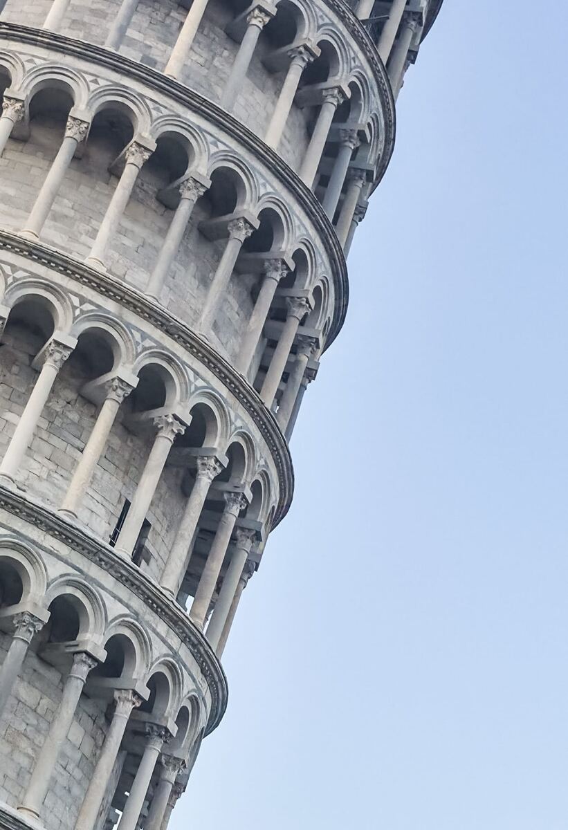 Een close-up van de scheve toren van Pisa, gefotografeerd schuin omhoog tegen een helderblauwe lucht. De toren toont meerdere galerijen met bogen en zuilen van lichtgekleurd steenwerk.