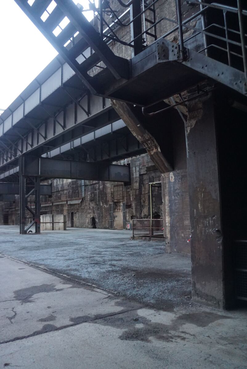 Een laag camerastandpunt toont de imposante stalen constructies en betonnen muren van een voormalig industrieel complex. Donkere metalen balken en platforms domineren de bovenzijde van het beeld, met een heldere, bijna witte lucht erboven. De voorgrond bestaat uit een mix van betonplaten en een grindpad, met sporen van vocht op de grond. De betonnen muren op de achtergrond zijn ruw en textureel.