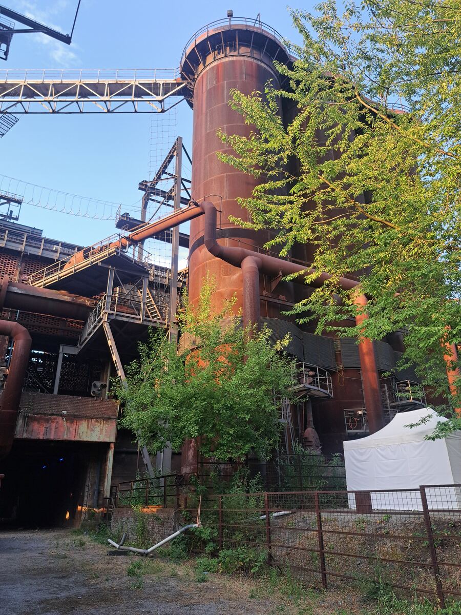 Een close-up van een grote roestbruine industriële installatie, waarschijnlijk een hoogoven, met pijpen, trappen en loopbruggen. Groene bomen en struiken groeien ervoor en ernaast. De lucht is blauw en helder. Rechtsonder staat een witte partytent. Links is een donkere doorgang zichtbaar.