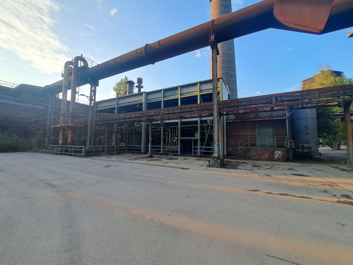 Een breed shot van een oud industrieel complex met grote roestige pijpleidingen die over een betonnen terrein lopen. Er zijn meerdere gebouwen zichtbaar met metalen constructies en een hoge schoorsteen op de achtergrond. De zon staat laag en werpt lange schaduwen, wat een warme gloed geeft aan de roestige oppervlakken. De lucht is helderblauw met enkele lichte wolken.