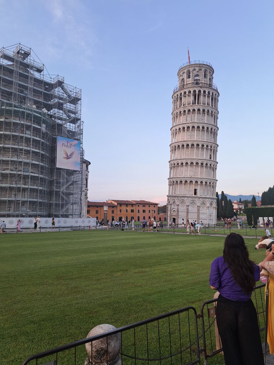 De foto toont het Piazza del Duomo in Pisa tijdens de avondschemering. Centraal staat de Scheve Toren van Pisa, een witmarmeren campanile met zijn kenmerkende helling en bogen. Links van de toren bevindt zich een groot gebouw, waarschijnlijk de Duomo of het Baptisterium, dat grotendeels bedekt is met steigers. Aan de steigers hangt een spandoek met de tekst 'Pace Peace' en een afbeelding van een duif. Op de voorgrond is een uitgestrekt groen gazon te zien, omzoomd door een hekwerk. Achter het he