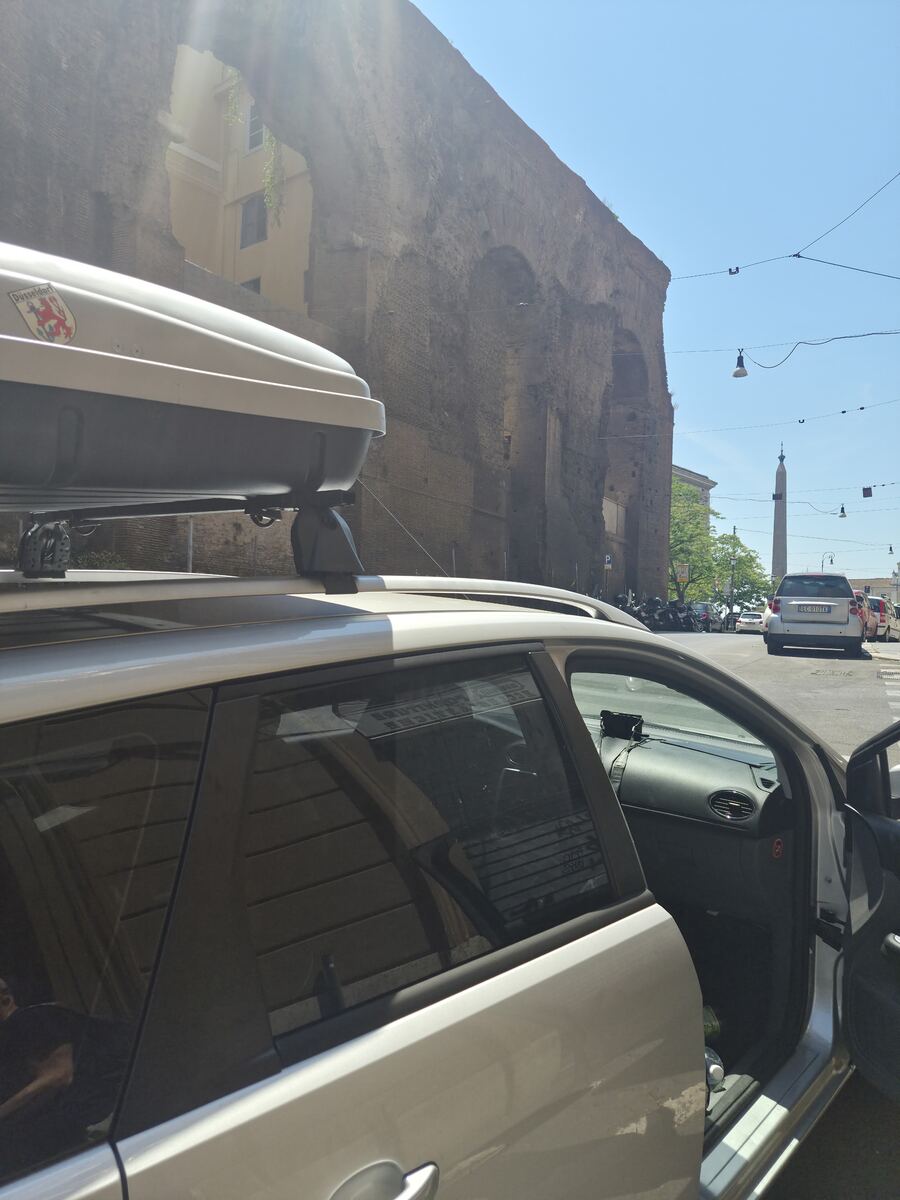 Een zilverkleurige Ford Focus stationwagon met een grijze dakkoffer staat geparkeerd langs een straat. Op de achtergrond is een grote, oude bakstenen muur met bogen te zien, mogelijk de overblijfselen van een aquaduct of een ander Romeins bouwwerk. De zon schijnt fel en er zijn bovengrondse elektriciteitskabels zichtbaar. Verderop in de straat staat een obelisk en zijn andere auto's en geparkeerde scooters zichtbaar. De passagiersdeur van de Ford Focus staat open.