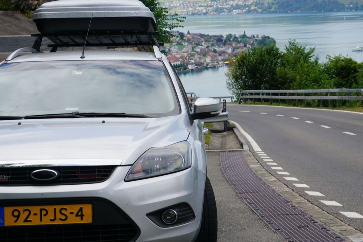 Een zilverkleurige Ford Focus stationwagen met een dakkoffer staat geparkeerd langs de kant van een geasfalteerde weg. Op de achtergrond is een groot meer te zien met huizen en bebouwing langs de oever en groene heuvels in de verte. De auto heeft een Nederlands kenteken (92-PJS-4).