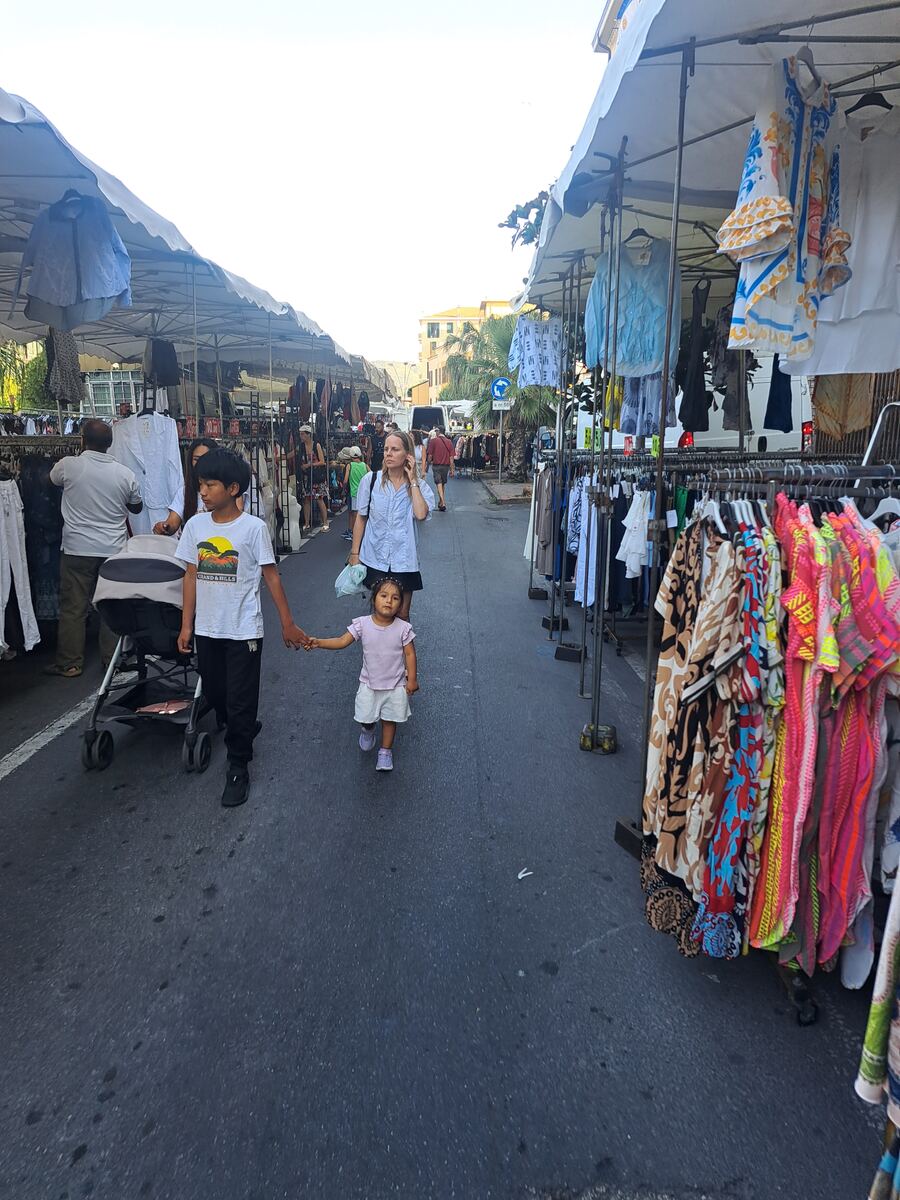 De foto toont een levendige buitenmarkt, waarschijnlijk een kledingmarkt, met rijen marktkramen onder witte luifels die zich uitstrekken langs een straat. Op de voorgrond lopen een jongen en een jong meisje hand in hand, met naast hen een kinderwagen. Rechts en links van hen staan rekken vol met diverse kledingstukken, waaronder kleurrijke jurken en shirts. Verschillende mensen zijn zichtbaar, sommigen bladeren door de kleding, anderen lopen door de gangpaden. Op de achtergrond zijn gebouwen, pa