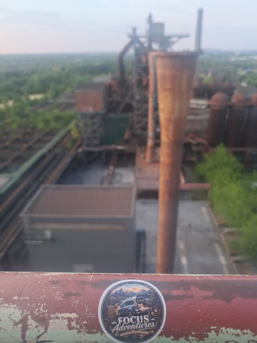 Een bovenaanzicht van een uitgestrekt industrieel complex met roestige structuren en gebouwen, deels begroeid met groen. Op de voorgrond is een roestige, groen en rood geverfde metalen reling zichtbaar met een ronde 'FOCUS Adventures' sticker. De achtergrond toont onscherpe, grote industriële installaties zoals hoogovens en pijpen onder een lichte avondlucht.