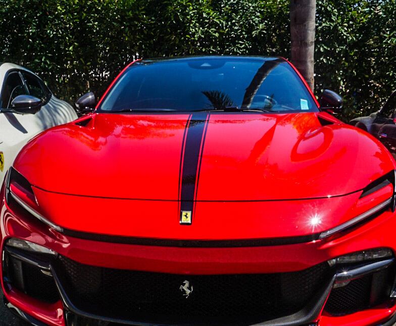 De voorkant van een felrode sportwagen van het merk Ferrari, met een zwarte racestreep over de motorkap en het dak. Het Ferrari-logo is zichtbaar op de motorkap en in de grille. Naast de rode auto is de zijkant van een witte auto, ook met een Ferrari-logo, deels zichtbaar. Op de achtergrond is groene beplanting te zien en een deel van een palmboomstam, alles onder helder zonlicht.