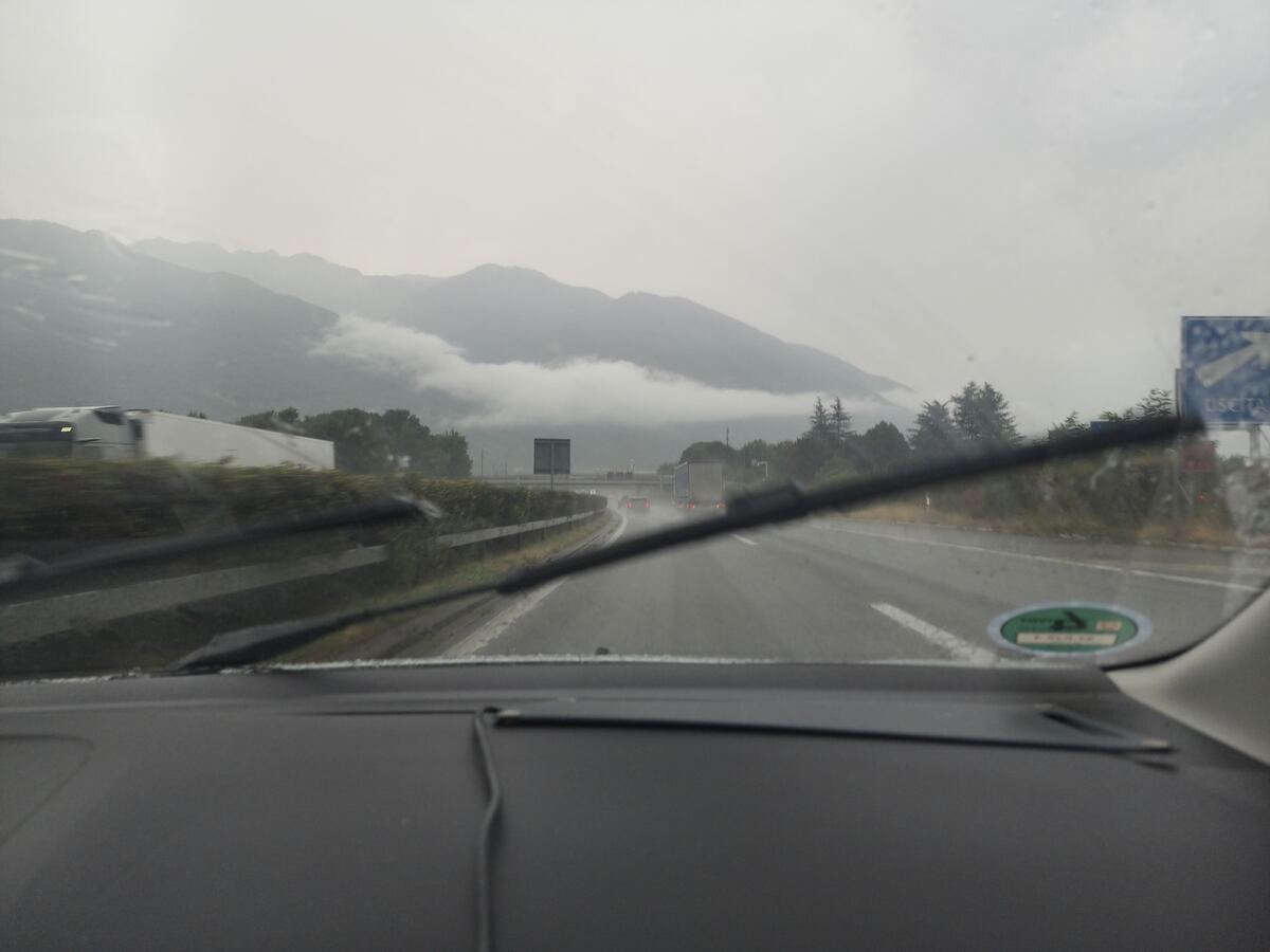 Een blik vanuit een auto door de voorruit op een natte snelweg met meerdere rijstroken. De ruitenwissers zijn zichtbaar in de onderste helft van het beeld, wat duidt op regen of recentelijke neerslag. In de verte zijn imposante bergen te zien, waarvan de toppen deels gehuld zijn in laaghangende wolken of mist. Langs de weg bevinden zich groene begroeiing, zoals struiken en bomen, en vangrails. Er rijden andere voertuigen, waaronder vrachtwagens en personenauto's, met brandende achterlichten. Aan