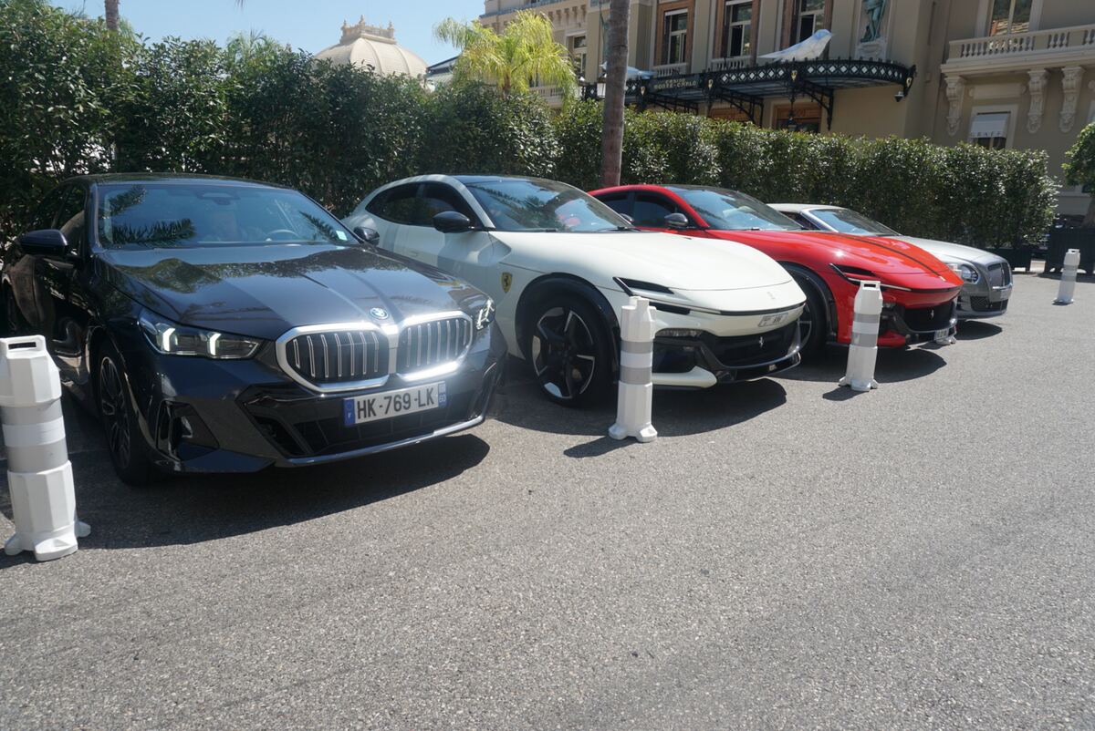 Een rij geparkeerde luxe auto's, waaronder een donkergrijze BMW, een witte Ferrari Purosangue, een rode Ferrari en een zilverkleurige Bentley, staat op een geasfalteerde ondergrond. Op de achtergrond is een statig gebouw met klassieke architectuur, balkons en dakkapellen te zien, omgeven door groene heggen en een palmboom. Witte afzetpaaltjes staan langs de weg. De zon schijnt helder en de naam 'MONTE-CARLO' is zichtbaar op de gevel van het gebouw.