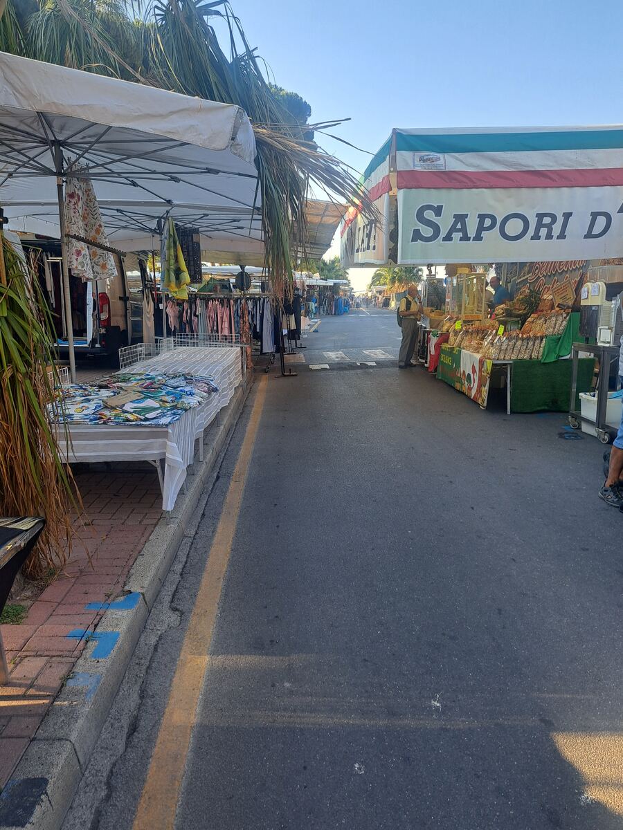 De foto toont een levendige straatmarkt onder een helderblauwe lucht, gefotografeerd vanuit een licht verhoogd perspectief. Aan weerszijden van de asfaltweg staan marktkramen met witte luifels, waaronder diverse goederen worden verkocht. Aan de linkerzijde zijn voornamelijk kleding en textiel te zien, uitgestald op rekken en tafels. Aan de rechterzijde staan kramen met voedingswaren, waaronder een kraam met een spandoek in de kleuren van de Italiaanse vlag en de tekst "SAPORI D'". Enkele mensen