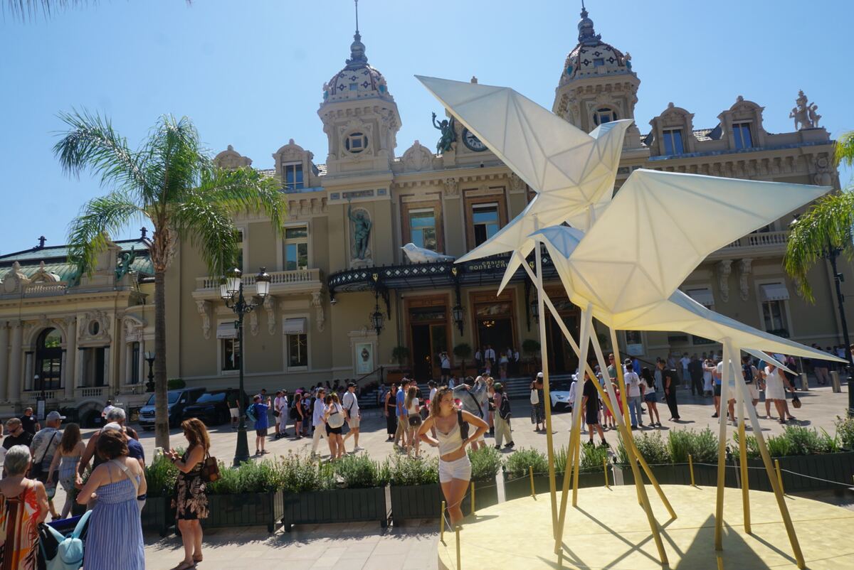 Een zonnige exterieurfoto van het Casino de Monte-Carlo in Monaco, een groot, sierlijk gebouw met koepels en beelden. Op de voorgrond staan twee grote, witte, geometrische sculpturen die op vogels lijken, op gouden sokkels. Er lopen veel mensen rond op het plein voor het gebouw. Een palmboom is zichtbaar aan de linkerkant onder een helderblauwe lucht.