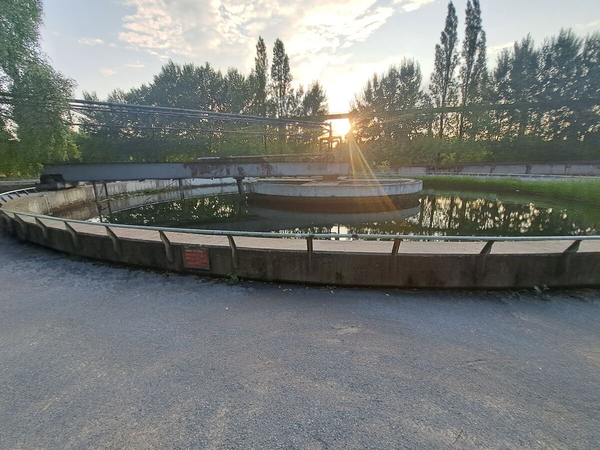 De foto toont een grote, ronde betonnen waterbekken met daarin een kleinere ronde structuur. Over het water loopt een metalen constructie met leidingen. Op de achtergrond staan bomen en de laagstaande zon schijnt door het gebladerte, wat reflecties in het water veroorzaakt. De voorgrond bestaat uit een geasfalteerd oppervlak en een betonnen rand met een metalen reling.