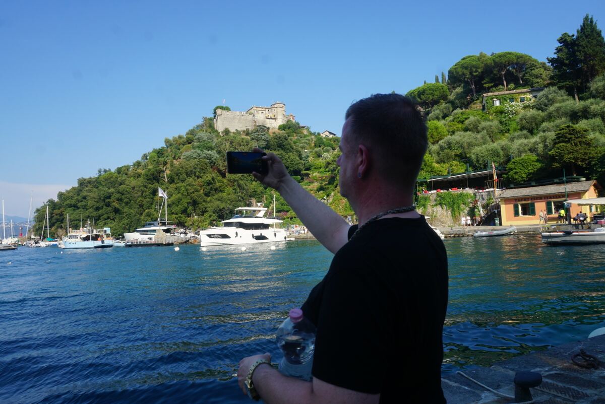 Een persoon met kort haar staat met de rug naar de camera gericht en maakt een foto met een smartphone van de haven van Portofino. Op de achtergrond is een heuvel te zien met weelderige groene begroeiing, diverse gebouwen, waaronder een kasteelachtig fort, en luxe jachten in het helderblauwe water. De lucht is onbewolkt en helderblauw.