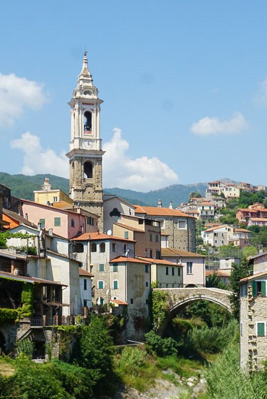 Een zonnig dorpsgezicht van Dolcedo in Ligurië, Italië. Centraal in de afbeelding staat een hoge, lichtgekleurde klokkentoren met een spits dak en een klok. Daaromheen liggen traditionele huizen met terracotta daken en gevels in diverse pasteltinten zoals roze, oker en wit, vaak voorzien van groene luiken. Een stenen boogbrug, deels begroeid met groen, overspant een droge bedding tussen de huizen. Op de achtergrond zijn groene, beboste heuvels zichtbaar onder een helderblauwe lucht met enkele wi