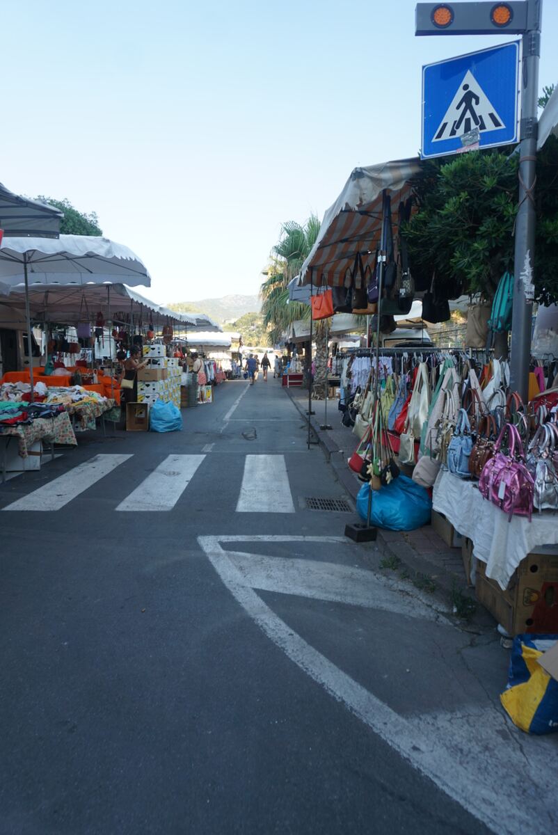 Een straatmarkt met aan weerszijden kramen onder afdaken. Op de kramen liggen en hangen diverse koopwaar, waaronder veel tassen en kleding. Op de voorgrond is een zebrapad zichtbaar, met een verkeersbord voor een oversteekplaats met knipperlichten ernaast. Achter de kramen zijn palmachtige bomen en in de verte heuvels onder een heldere lucht te zien. Enkele mensen lopen verderop in de straat.