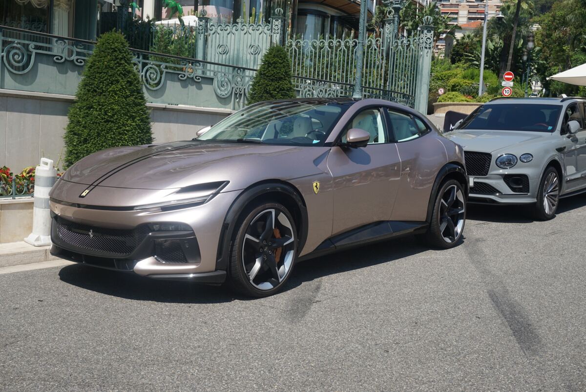 Op de foto is een lichtbruine Ferrari Purosangue te zien, geparkeerd op een geasfalteerde weg. Achter de Ferrari staat een grijze Bentley Bentayga geparkeerd. Op de achtergrond is een gebouw met een sierlijk hekwerk en groene, kegelvormige struiken zichtbaar. Rechtsboven in beeld hangt een verkeersbord met een rood omrande cirkel en het getal 10 erin. De zon schijnt helder en werpt duidelijke schaduwen op de weg.