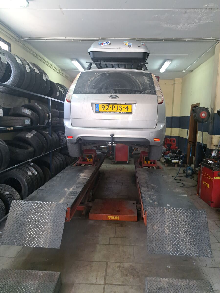 Een zilverkleurige Ford Focus stationwagon met een dakkoffer staat op een autolift in een garage. De achterkant van de auto, inclusief een Nederlands kenteken (92-PJS-4) en een stoffige achterruit, is prominent in beeld. Aan de linkerzijde van de garage zijn hoge stellingen gevuld met diverse autobanden. Aan de rechterkant staan verschillende gereedschappen en garage-apparatuur, waaronder een rode kast met de tekst 'STAR' en een haspel. De ruimte wordt verlicht door tl-lampen aan het plafond en