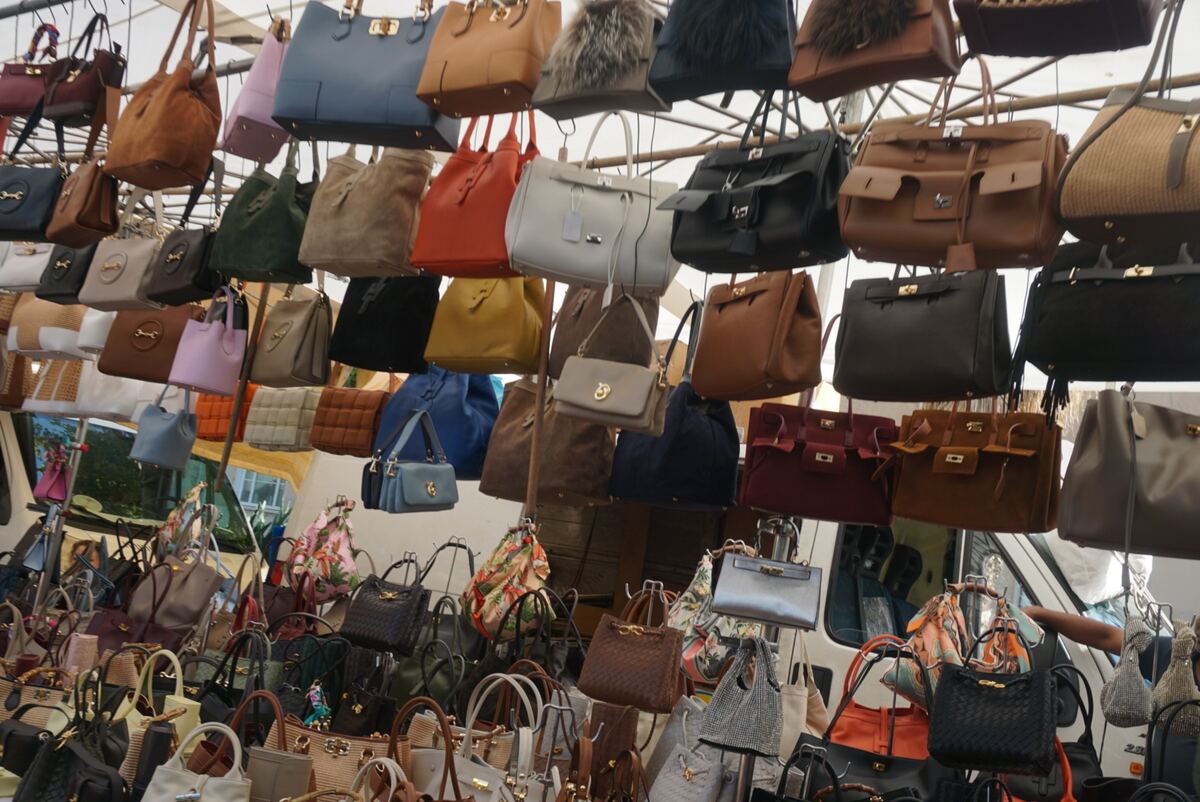 Een overzicht van een marktkraam vol met een grote verscheidenheid aan handtassen in verschillende kleuren, maten en stijlen. De tassen hangen dicht op elkaar aan metalen rekken, zowel hoog als lager, en vullen het grootste deel van het beeld. We zien tassen in leerachtige materialen, suède en geweven stoffen, met diverse sluitingen en details. De kleuren variëren van neutrale tinten zoals bruin, beige en grijs tot opvallende kleuren zoals rood, oranje, blauw, geel en paars. Op de achtergrond is