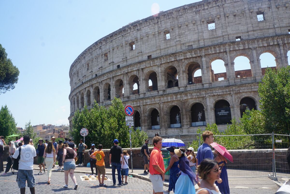 Een groots deel van de buitenmuur van het Colosseum is zichtbaar onder een helderblauwe lucht. De stenen constructie met meerdere lagen bogen domineert het beeld. Op de voorgrond en langs de zijkant van de weg lopen veel mensen, sommigen met parasols. Er staan groene bomen en struiken langs de rand, en een verkeersbord is zichtbaar. Binnen enkele bogen zijn tentoonstellingsstukken te zien en een spandoek met 'the Colosseum tells its story'.
