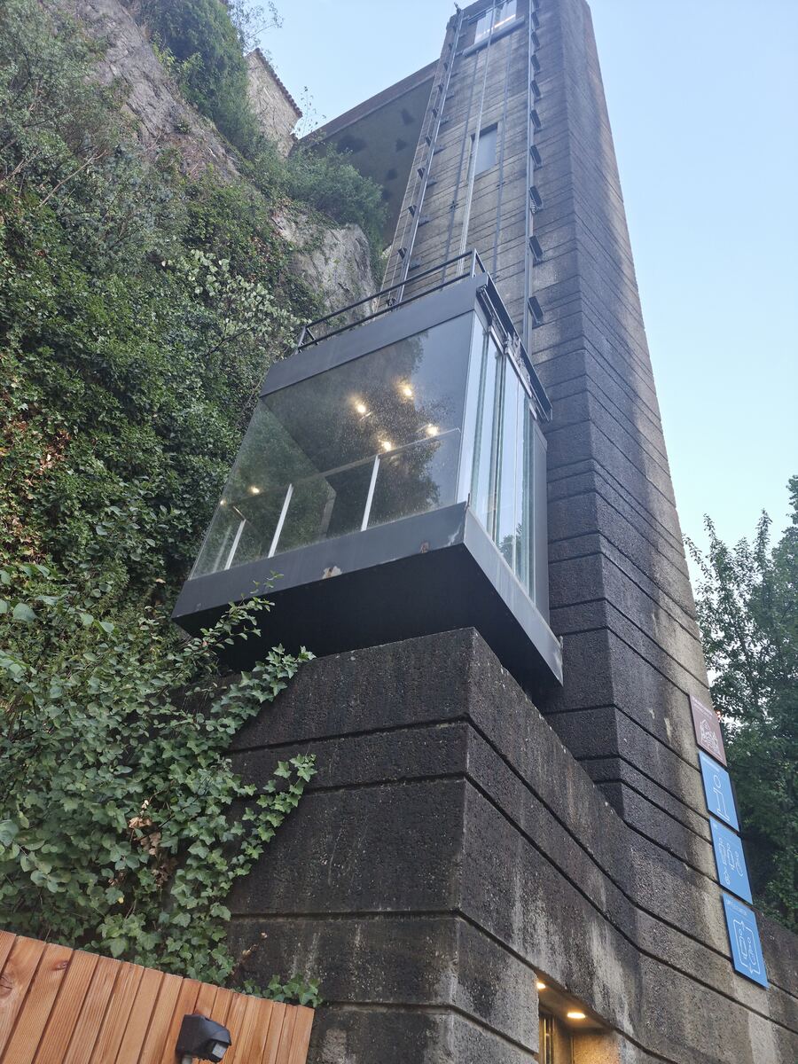 Een moderne glazen liftcabine, verlicht van binnenuit, is bevestigd aan een hoge betonnen toren die tegen een steile, begroeide rotswand is gebouwd. De toren heeft horizontale groeven en aan de zijkant zijn enkele blauwe bordjes met pictogrammen zichtbaar. Boven de rotswand is een stukje van een gebouw met een rood dak te zien, en daarboven een helderblauwe avondlucht. Onderaan de foto is een houten schutting en een bewegingssensor te zien.