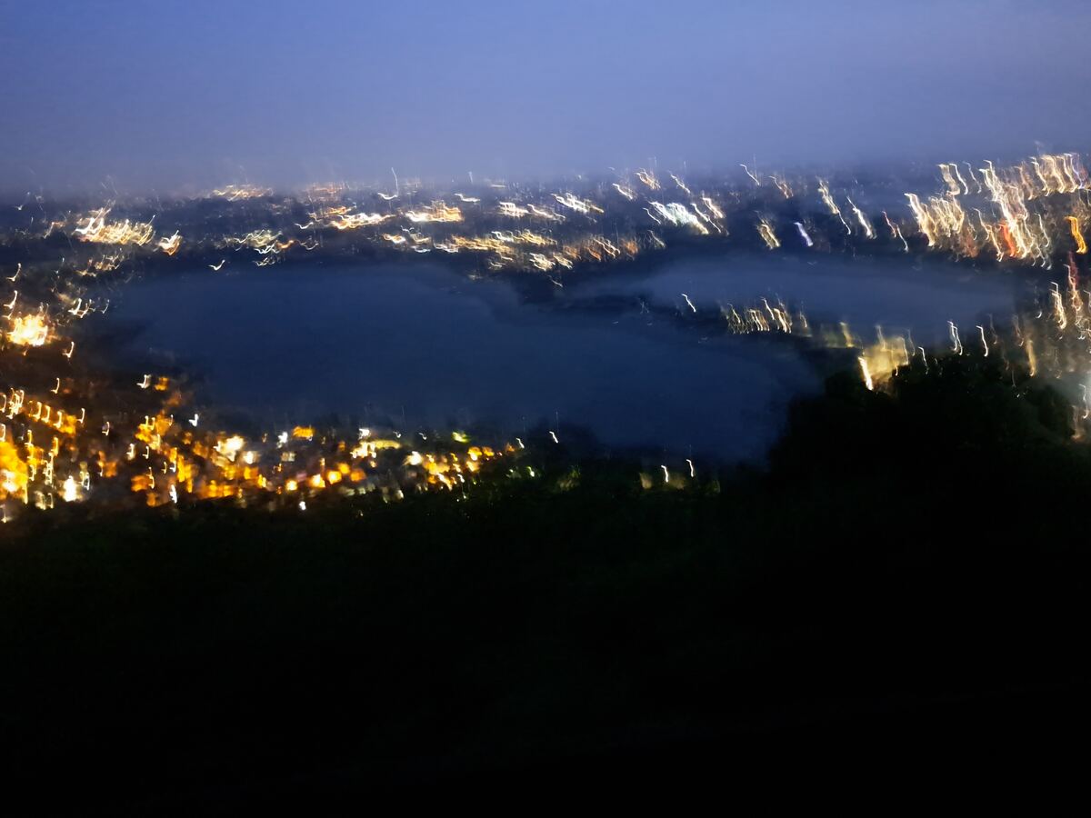 Een wazige nachtfoto toont een panoramisch uitzicht over een donkerblauw waterlichaam, waarschijnlijk een meer, omringd door talloze onscherpe, langgerekte lichtstrepen van een stad of dorp. De lichtjes variëren in kleur van geel en oranje tot wit, en strekken zich uit over een groot deel van het landschap. De horizon is een donkerblauwe tot paarse lucht, en op de voorgrond zijn donkere, silhouetachtige contouren van vegetatie of heuvels zichtbaar. De onscherpte van de lichten suggereert bewegin