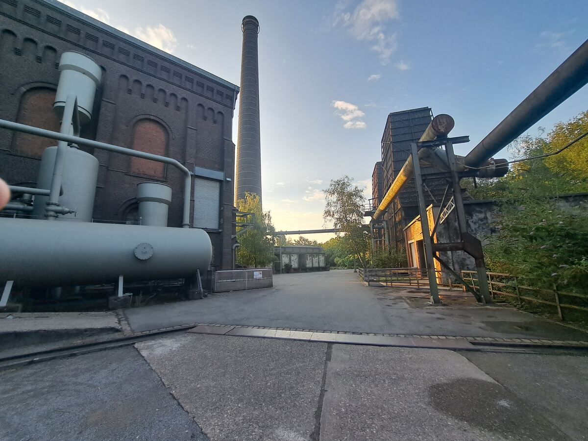 Een breed shot van een oud industrieel complex met een hoge schoorsteen in het midden. Links staat een groot bakstenen gebouw met ronde raamopeningen en daartegenaan staan grote grijze tanks en leidingen. Rechts zijn meer metalen constructies en leidingen zichtbaar, met een kleiner bakstenen gebouw eronder. Overal tussen de structuren door is groen te zien, en de lucht is lichtblauw met enkele witte wolken, verlicht door de ondergaande zon die een warme gloed geeft op sommige delen van de gebouw
