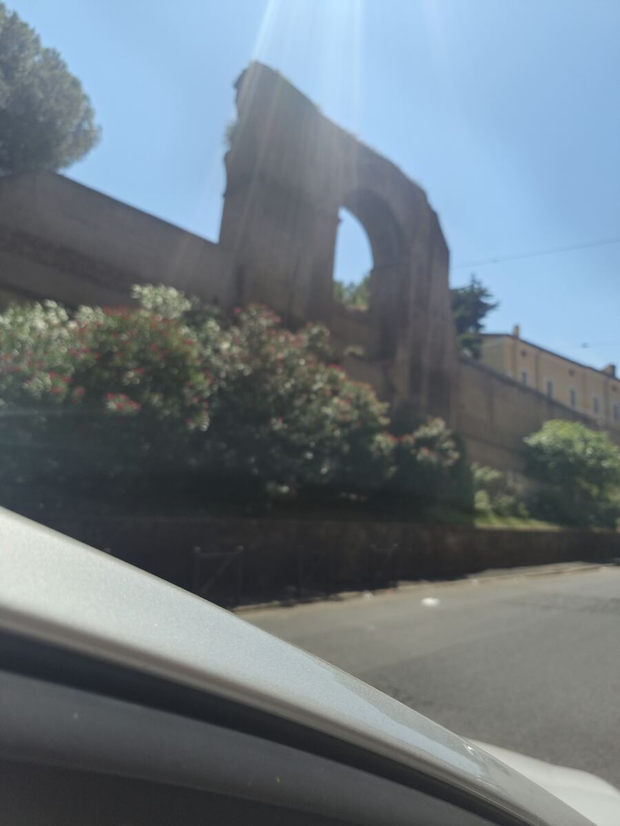 Een enigszins onscherpe foto genomen vanuit een auto, met een deel van de zilverkleurige carrosserie linksonder in beeld. Op de achtergrond is een oud, deels vergaan stenen bouwwerk te zien, mogelijk een aquaduct of een oude muur met een boogopening. Er zijn groene struiken met roodachtige bloemen langs het bouwwerk. De lucht is helderblauw en er is een felle zonnestraal zichtbaar in het midden van de foto. Rechtsachter het bouwwerk is een lichtgeel gebouw met ramen te onderscheiden. Een deel va