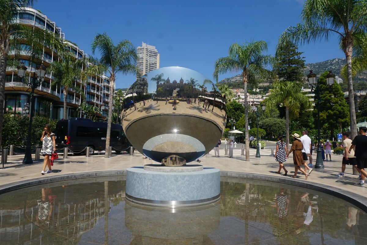Een grote, glimmende bolsculptuur staat in een ondiepe waterpartij op een zonnig plein in Monaco. De bol reflecteert de omgeving, waaronder een statig gebouw met koepels, palmbomen en mensen die voorbijlopen. Op de achtergrond zijn moderne gebouwen met balkons en groene heuvels te zien onder een helderblauwe lucht.