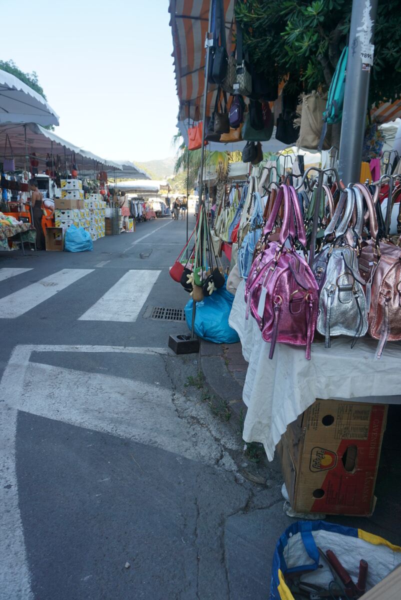 Een brede straat met een levendige markt onder gestreepte luifels. Aan de rechterkant van de foto zijn glimmende handtassen in roze, zilver en roségoud te zien, uitgestald op een tafel. Aan de linkerkant staan kramen met dozen vol fruit, mogelijk bananen, en kleding. Op de voorgrond is een zebraoversteek zichtbaar op het asfalt. In de verte lopen mensen tussen de marktkramen en zijn heuvels onder een heldere, lichte lucht te zien. Een lantaarnpaal met een nummer '7896' is rechts in beeld.