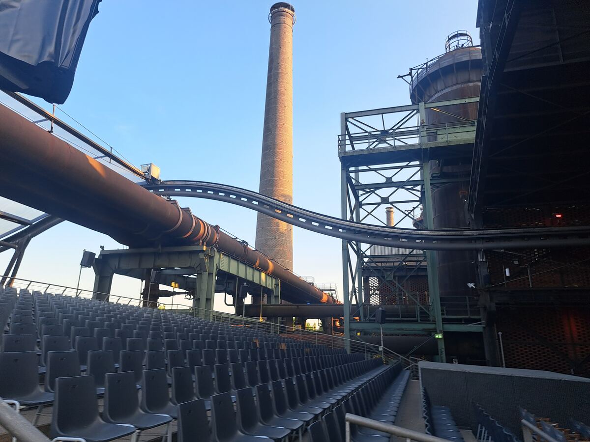 Een weids zicht op een voormalig industrieel complex met een hoge bakstenen schoorsteen in het midden. Grote roestbruine pijpleidingen en metalen constructies domineren de voorgrond en middengrond. Rechts is een deel van een grote ronde tank of toren zichtbaar. Op de voorgrond zijn rijen lege, donkere stoelen van een amfitheater te zien. De lucht is helderblauw met een zachte avondgloed die de structuren raakt.