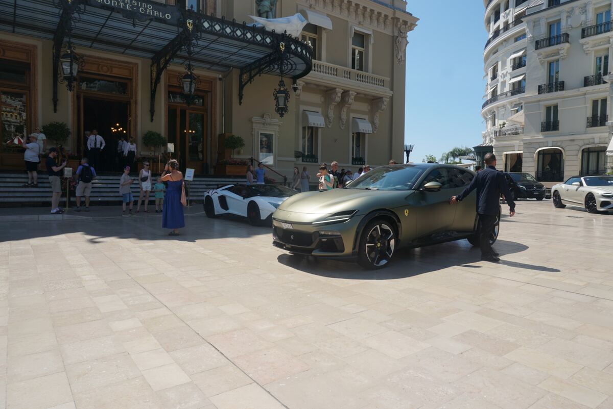 Een zonnige straat voor het Casino de Monte-Carlo, een groot, klassiek gebouw met een sierlijke luifel boven de ingang. Op de voorgrond staat een matte olijfgroene Ferrari Purosangue, en verderop is een witte Lamborghini Aventador zichtbaar. Meerdere mensen lopen op de lichtgekleurde tegelbestrating, sommigen kijken naar de auto's of gaan het casino in. Op de achtergrond zijn meer elegante gebouwen te zien onder een helderblauwe lucht.