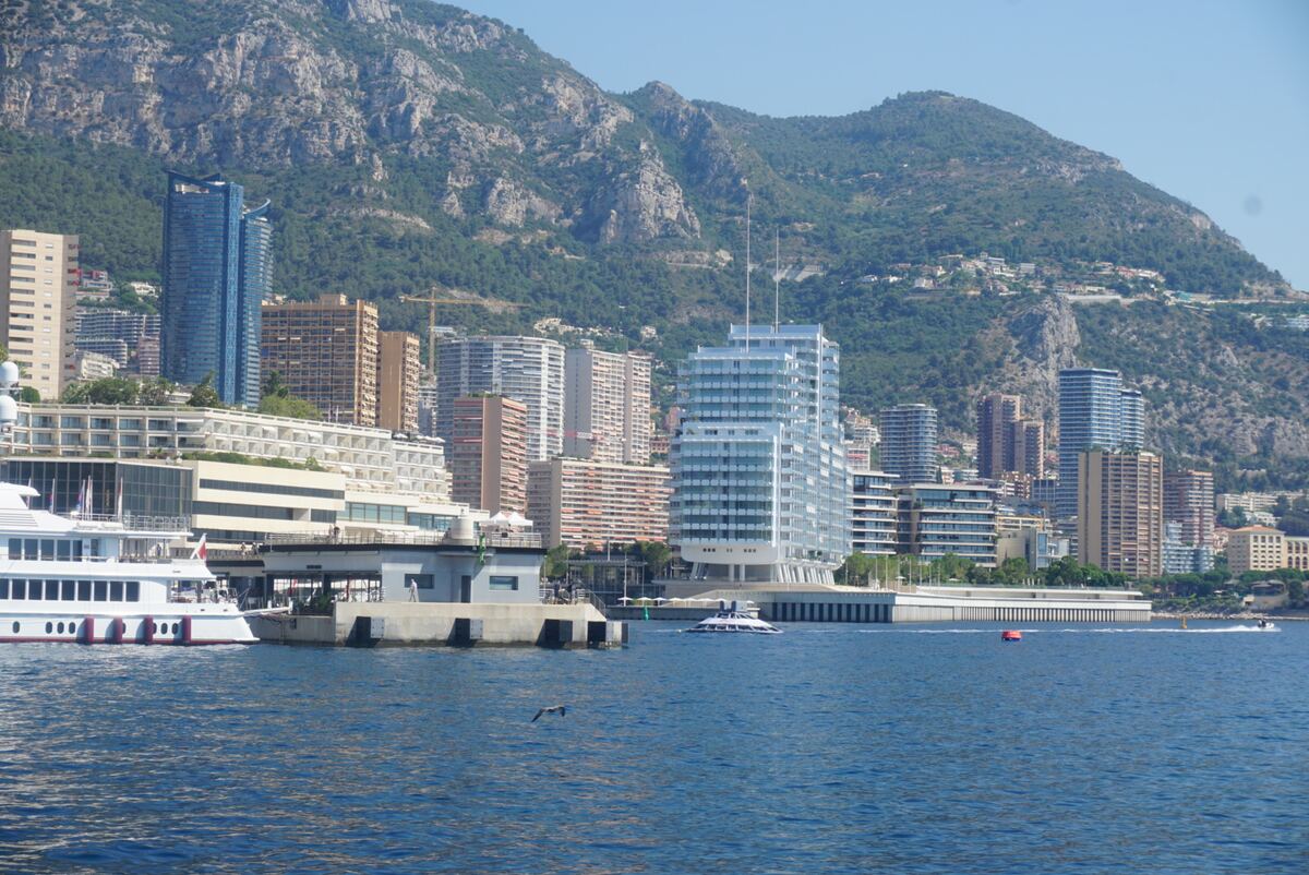 Een brede blik op de kustlijn van Monaco met de haven op de voorgrond. Het water is diepblauw en diverse boten, waaronder een grote witte jacht, liggen in het water of varen. Op de achtergrond rijzen veel hoge gebouwen en appartementencomplexen op tegen een steile, begroeide bergwand. De lucht is helderblauw en er vliegt een vogel laag over het water.