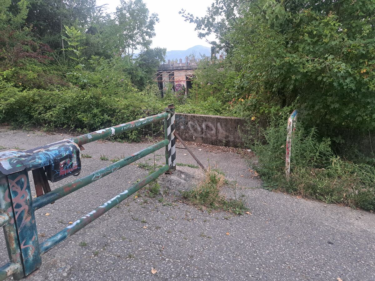 De foto toont een deels geopende, groen geverfde metalen slagboom in de voorgrond, rijkelijk voorzien van graffiti. De slagboom heeft drie horizontale spijlen en een verticale paal met zwart-witte strepen. Het pad is geasfalteerd met hier en daar onkruid. Achter de slagboom is een betonnen muur te zien, eveneens met graffiti. Daarachter bevindt zich een dichtbegroeid gebied met bomen en struiken. Door een opening in het groen is een deel van een gebouw zichtbaar met een decoratieve, mogelijk gek
