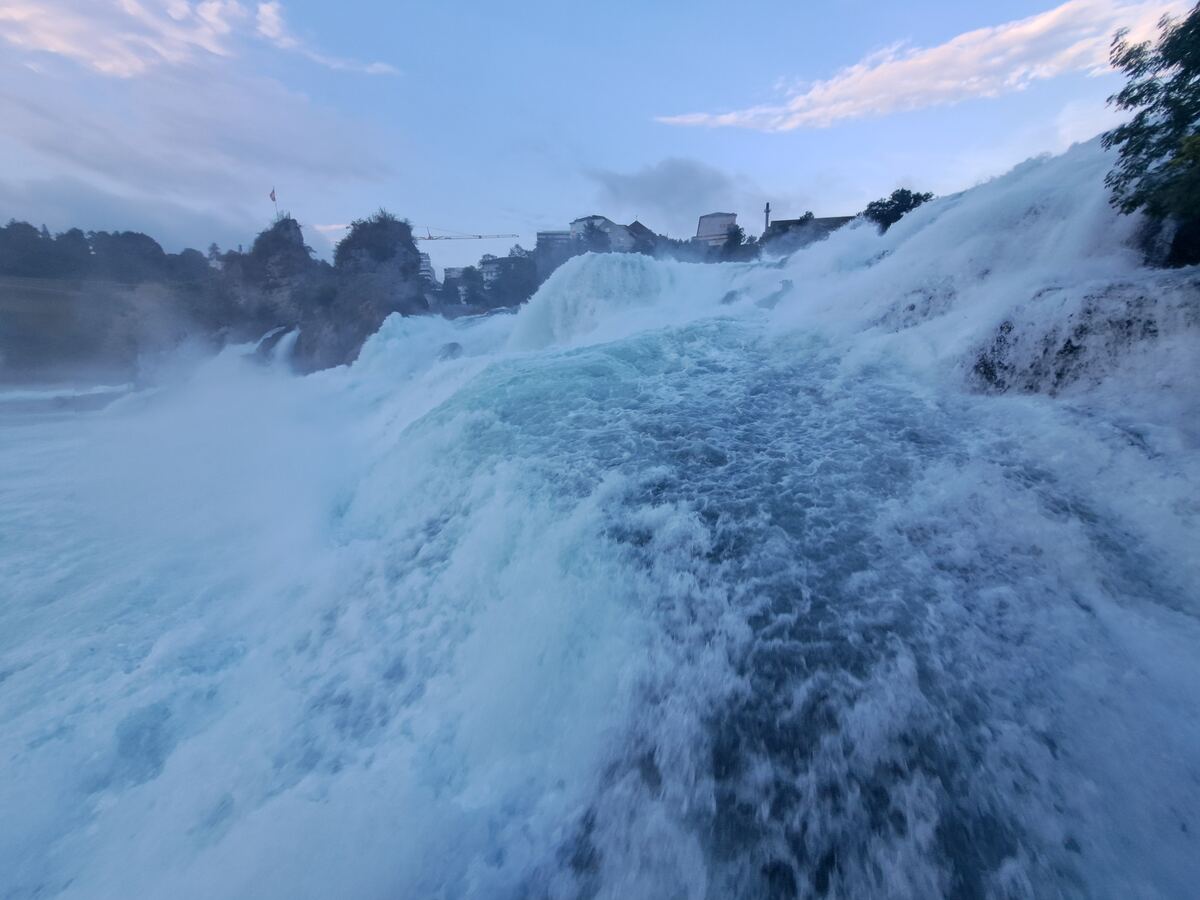 De foto toont de Rijnwaterval vanuit een laag perspectief, met een grote hoeveelheid turbulent, wit water dat van linksboven naar rechtsonder stroomt. Op de achtergrond zijn rotsachtige oevers met bomen en enkele gebouwen zichtbaar. Aan de linkerkant, op een rotspunt, wappert een rode vlag. Een hijskraan is in de verte te zien boven de gebouwen. De lucht is deels bewolkt en kleurt zachtblauw, wat duidt op de late avond.