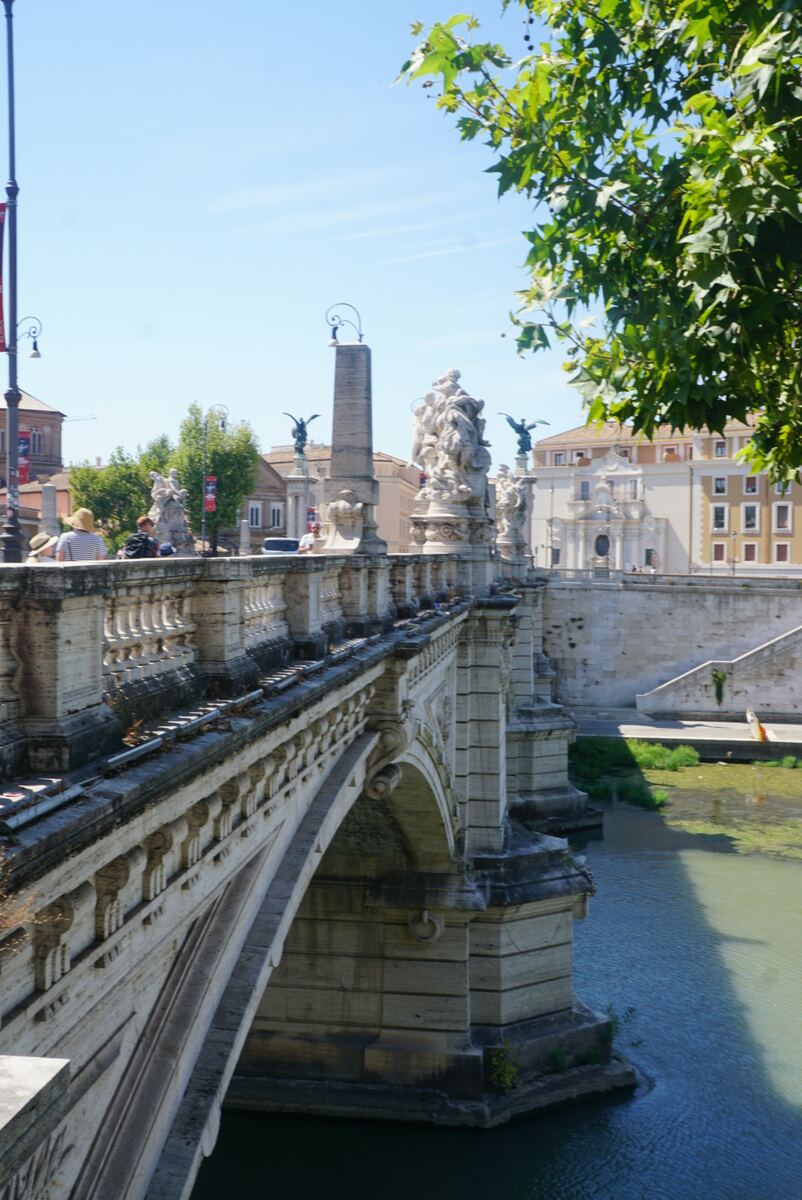 Een foto van een imposante stenen brug met rijk versierde balustrades en beelden aan weerszijden. Onder de bogen van de brug stroomt water, deels bedekt met groen. Aan de overkant van het water zijn gebouwen te zien, waaronder een met een klassieke gevel. De lucht is helderblauw en er zijn bomen met groene bladeren zichtbaar, deels rechtsboven in beeld. Enkele mensen lopen over de brug.