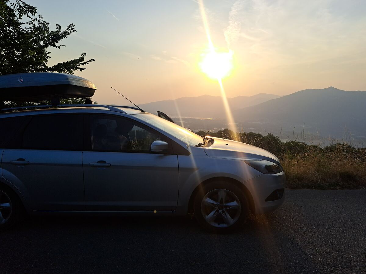 De foto toont een zilverkleurige stationwagen met een dakkoffer op het dak, geparkeerd aan de rechterkant van een geasfalteerde weg. De auto is gedeeltelijk in de schaduw, maar de motorkap en voorruit worden beschenen door de felle, laaghangende zon. Op de achtergrond strekt zich een berglandschap uit met meerdere lagen heuvels en bergen die vervagen in de verte. In de vallei onder de bergen zijn contouren van bebouwing en mogelijk een waterlichaam zichtbaar, reflecterend in het zonlicht. Aan de