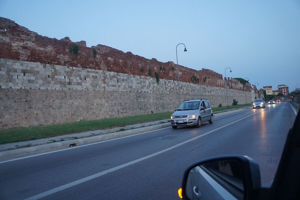 Een foto genomen vanuit een rijdende auto, met een lange, oude stadsmuur aan de linkerkant. De muur bestaat uit verschillende lagen steen en baksteen en is deels begroeid met kleine planten. Rechts van de muur loopt een weg met meerdere auto's, waarvan de koplampen branden in de avondschemering. De lucht is schemerig blauw. Een autospiegel is rechtsonder in beeld zichtbaar, met een oranje knipperlichtreflectie.
