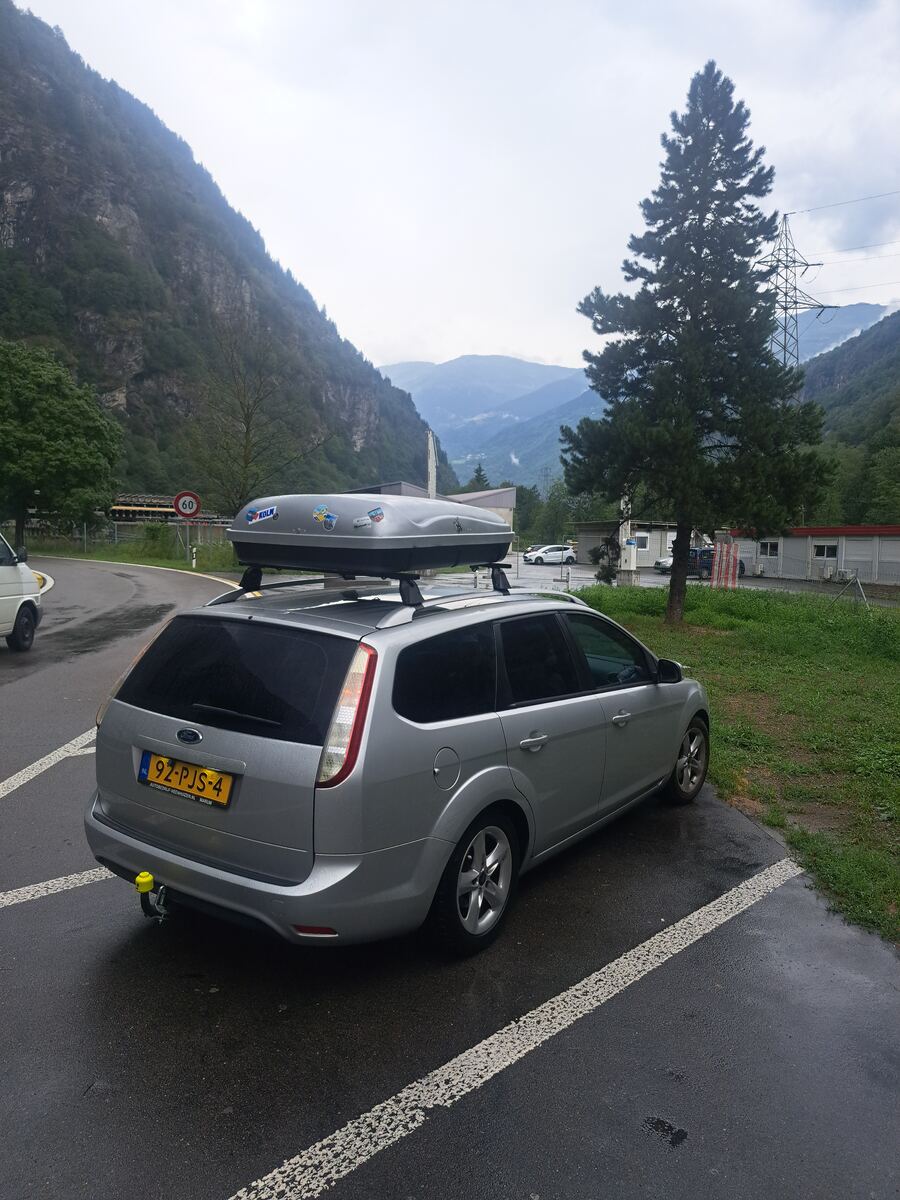 Een zilverkleurige Ford Focus stationwagon met een grijze dakbox staat geparkeerd op een nat asfaltterrein. De auto heeft een Nederlands kenteken '92-PJS-4'. Op de achtergrond zijn steile, beboste bergen te zien onder een bewolkte, grijze hemel. Rechts van de auto staat een hoge naaldboom en zijn enkele lage, functionele gebouwen zichtbaar. Aan de linkerzijde van de weg is een snelheidsbord met '60' te zien, evenals een deel van een witte bestelwagen. De dakbox is versierd met meerdere stickers,