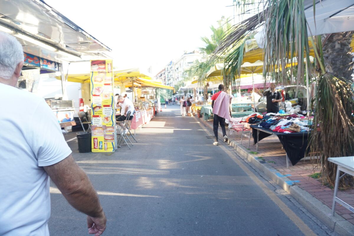Een straatmarkt in Ventimiglia met diverse kraampjes onder gele parasols en luifels. Aan de linkerkant staan foodtrucks en eettentjes, aan de rechterkant liggen kleding en andere koopwaar uitgestald. Enkele palmbomen zijn zichtbaar. Er zijn mensen op de markt, waaronder een persoon op de voorgrond met de rug naar de camera.