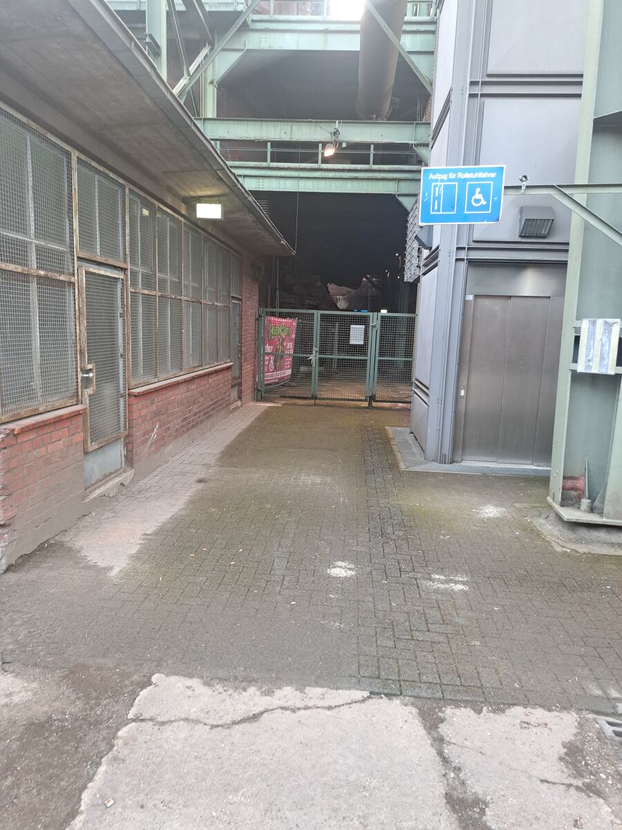 Een geplaveid pad loopt tussen een bakstenen gebouw met ramen voorzien van metalen gaas en een stalen constructie met een lift. Boven het pad hangen industriële balken en buizen. Op de stalen constructie is een blauw bord zichtbaar met pictogrammen voor een lift en een rolstoelgebruiker, met de tekst 'Aufzug für Rollstuhlfahrer'. De omgeving is donker, wat duidt op de avond, met enkele lichtpunten van lampen.