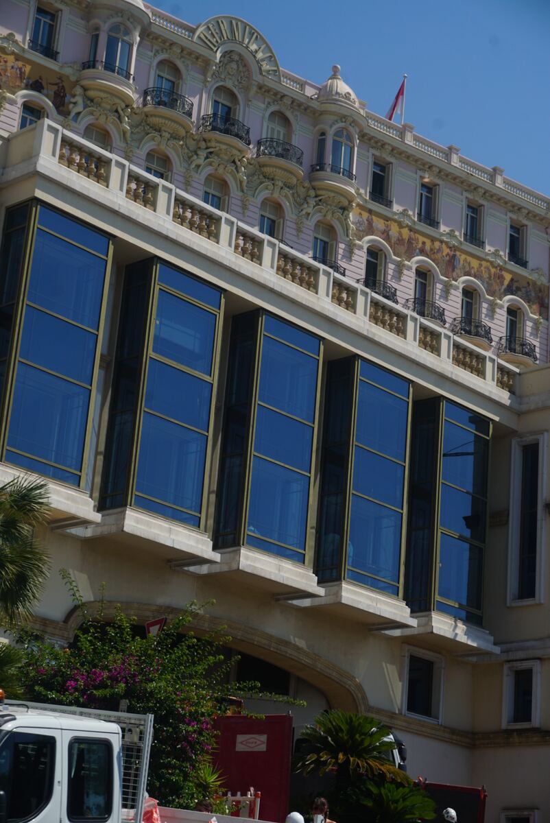 Een close-up van de gevel van een groot, luxueus gebouw. De bovenste verdiepingen zijn lichtroze met veel ornamenten, smeedijzeren balkons en muurschilderingen. Op het dak wappert een rood-witte vlag. De onderste verdiepingen bestaan uit moderne, blauw getinte glazen panelen. Enkele palmbomen en struiken zijn zichtbaar aan de onderkant van de foto, samen met een deel van een wit voertuig.