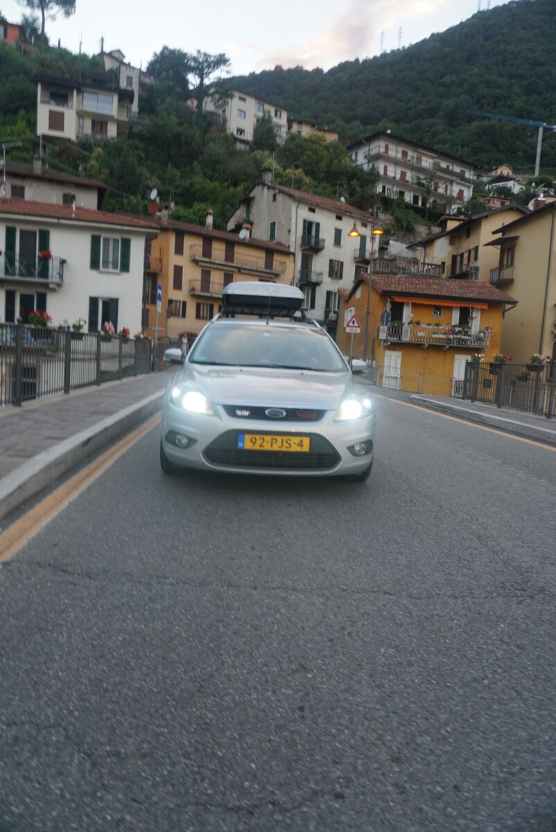 Een zilverkleurige Ford Focus stationwagen met een dakkoffer rijdt op een asfaltweg. De koplampen zijn aan. Aan weerszijden van de weg staan huizen, waarvan vele op een steile, beboste heuvel zijn gebouwd. Op de voorgrond links is een metalen hek met bloembakken zichtbaar. De lucht is schemerig en de straatverlichting is aan.