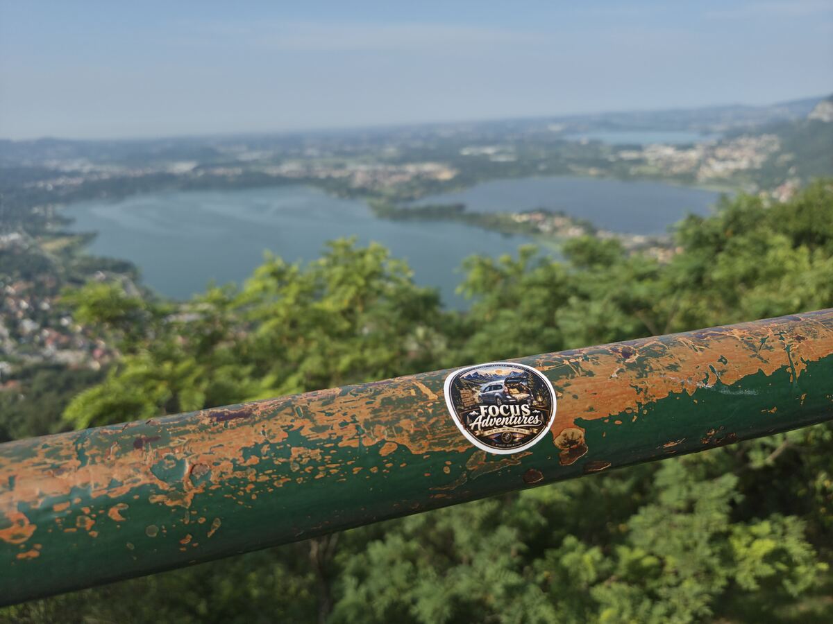 Op de voorgrond is een verweerde, groen geverfde metalen reling te zien, waarvan de verf afbladdert en een roestkleurige onderlaag onthult. Op de reling is een ronde sticker geplakt met de tekst 'Focus Adventures' en afbeeldingen van een terreinwagen en bergen. Achter de reling bevindt zich weelderig groen gebladerte. Op de achtergrond strekt zich een groot, blauwgroen meer uit, omzoomd door wat lijkt op bebouwing en heuvelachtig landschap onder een helderblauwe lucht.